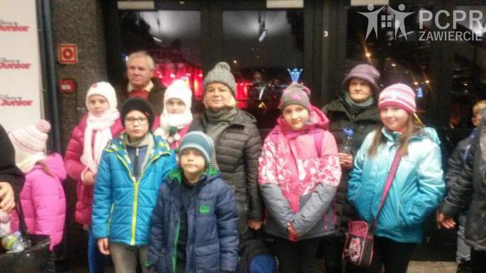 Zdjęcie: Grupa dzieci i dorosłych w kurtkach i czapkach stoi przed szklanymi drzwiami